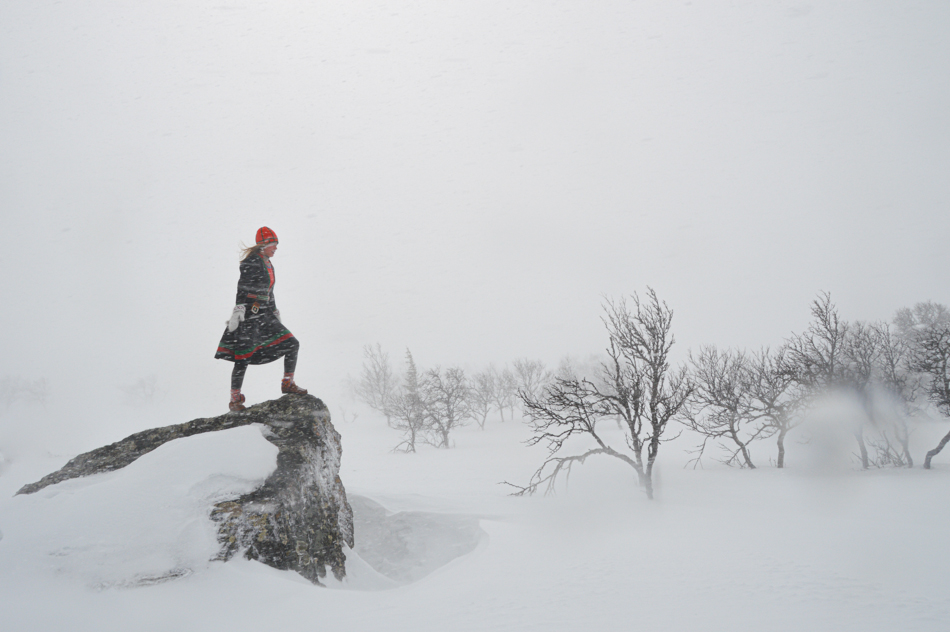Kvinna i kolt står på en stor sten på kalfjället. Det är vinter och motvind.