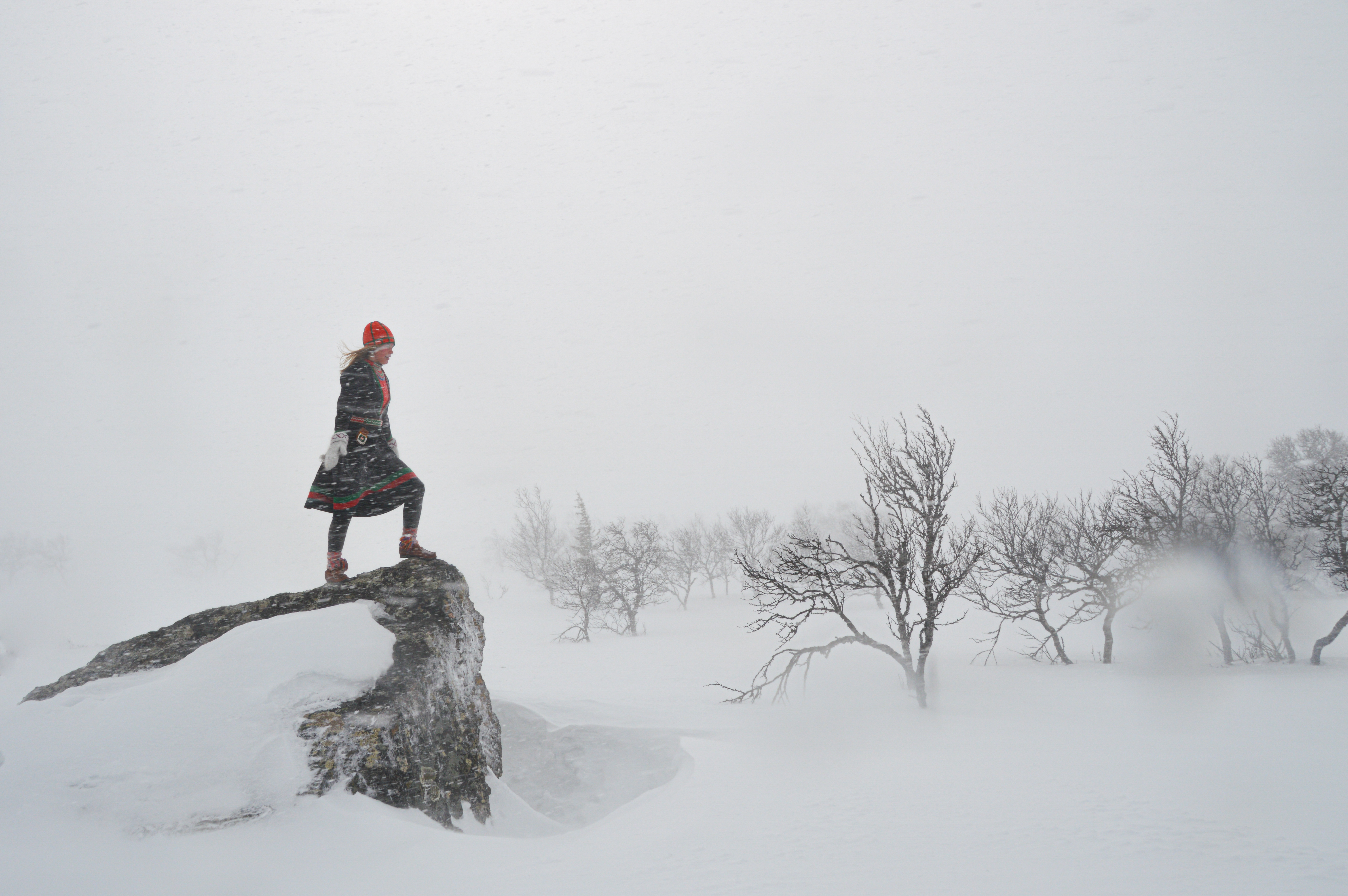 En ung kvinna i kolt stor på en sten på kalfjället mitt i en snöstorm.